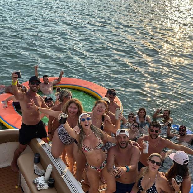 A large group crowded on the stern with drinks, an orange floating pool behind them on sunlit water