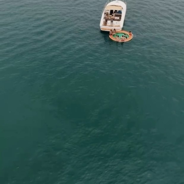 A single boat far out on flat green water, seen from high above, with a float tied alongside